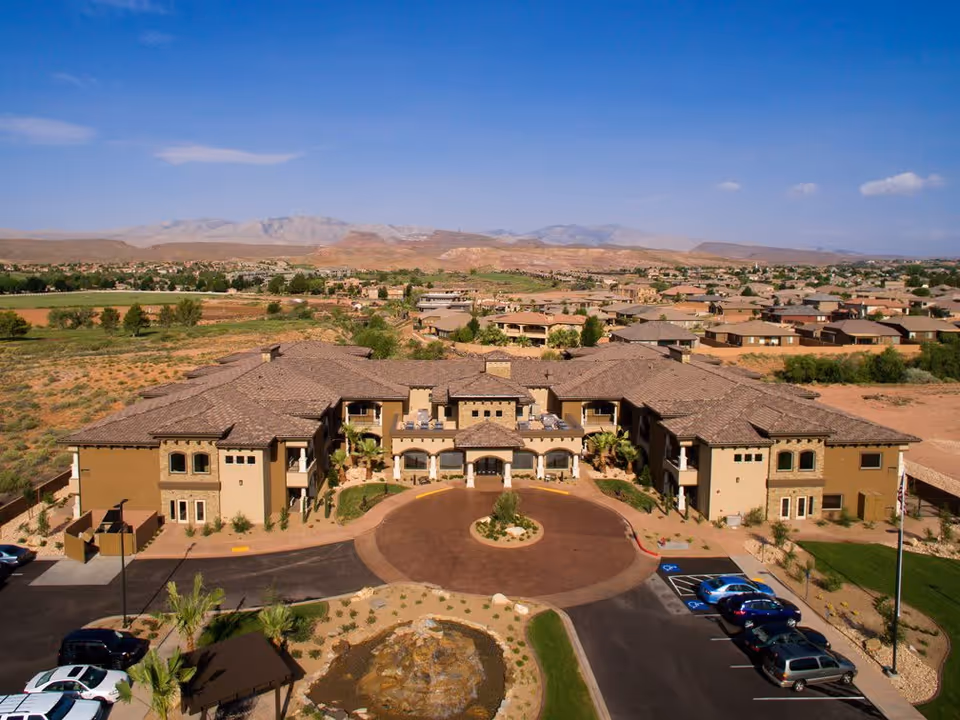 Aerial view of a large, single-story senior living facility with a circular driveway and landscaped entrance, surrounded by a desert landscape and distant mountains under a clear blue sky.