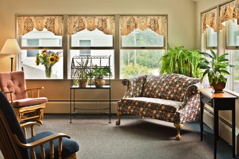 A cozy living room area with a floral patterned sofa, a pink cushioned rocking chair, and a blue cushioned wooden chair. There are several potted plants on a metal plant stand and a side table. The room has large windows with floral valances, letting in natural light and showing a view of greenery outside.