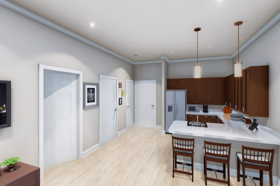 Interior view of a modern kitchen with wooden cabinets, white countertops, and three wooden bar stools at a breakfast bar. The kitchen includes a refrigerator, microwave, coffee maker, and toaster. The room has light-colored wood flooring and beige walls with white trim. There are three closed white doors along one wall, and framed pictures hanging between them. Two pendant lights hang over the breakfast bar.