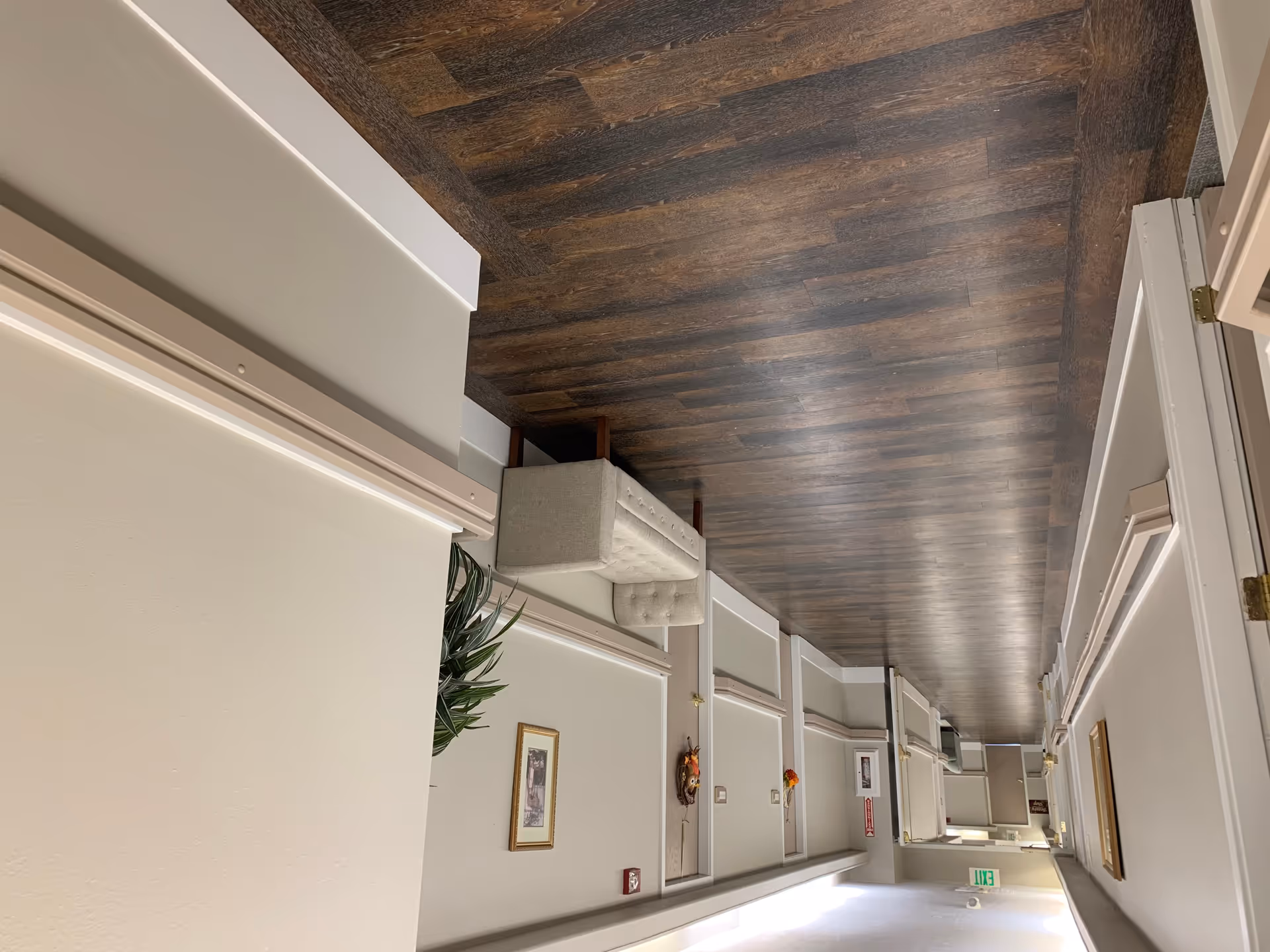 A clean and well-lit hallway in an assisted living facility with wood-patterned flooring, light-colored walls, a small upholstered bench, framed artwork, and an exit sign at the far end.