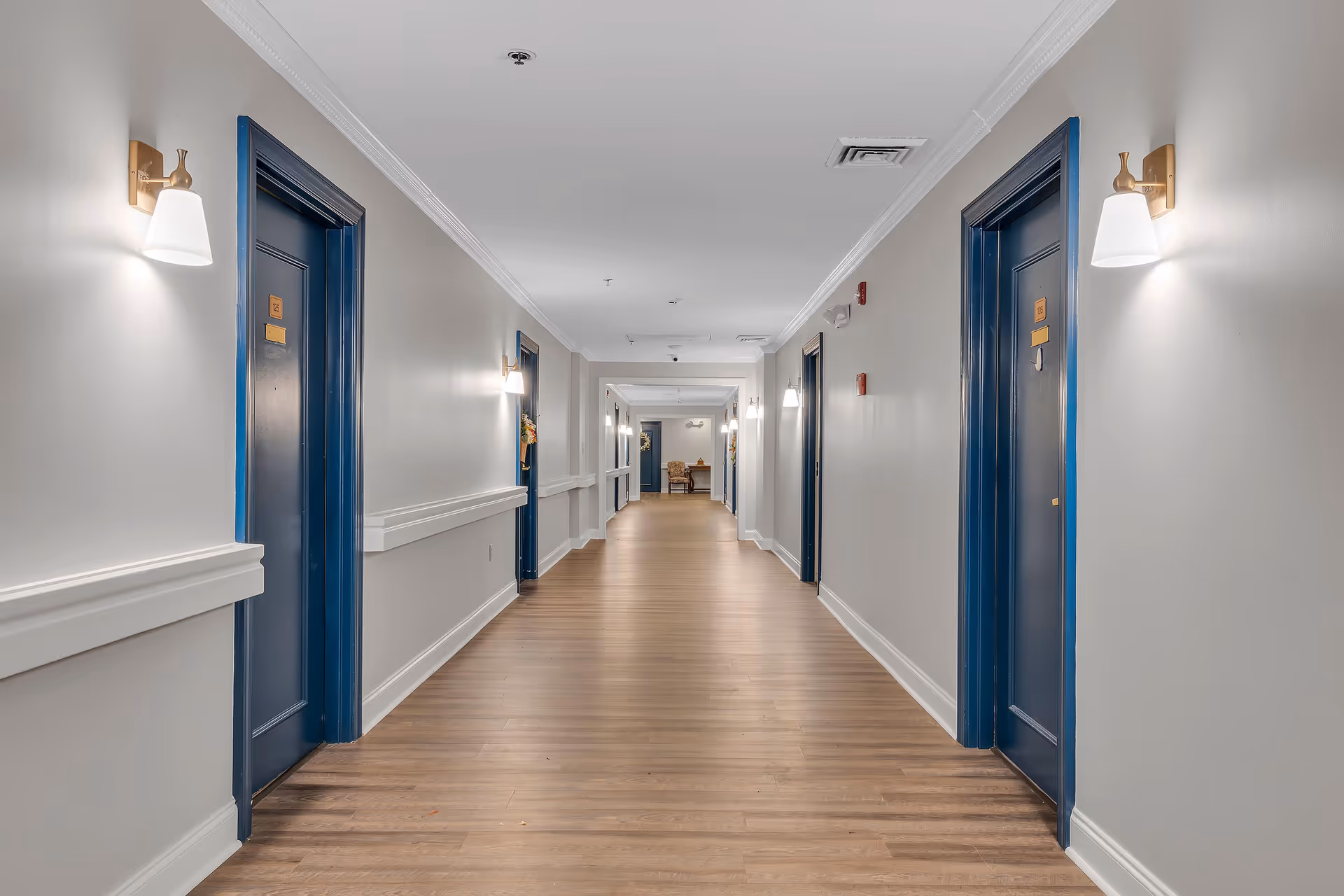 A long, well-lit hallway in a senior living facility with wooden flooring, white walls, and blue door frames. Wall-mounted light fixtures are evenly spaced along the corridor, and several closed doors line both sides of the hallway.