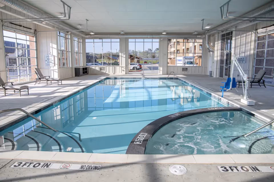 Indoor swimming pool area with large windows allowing natural light to fill the space. The pool has steps leading into the water and a connected circular hot tub with bubbling water. Several lounge chairs are placed around the pool deck. The room has a high ceiling with exposed ductwork and a clean, modern design.