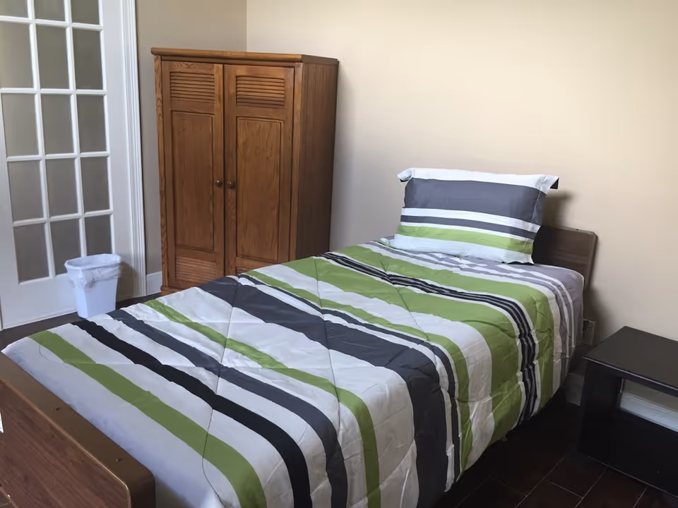 Single bed with green, gray, black and white striped bedding beside a wooden wardrobe and glass-paneled door in a small bedroom.