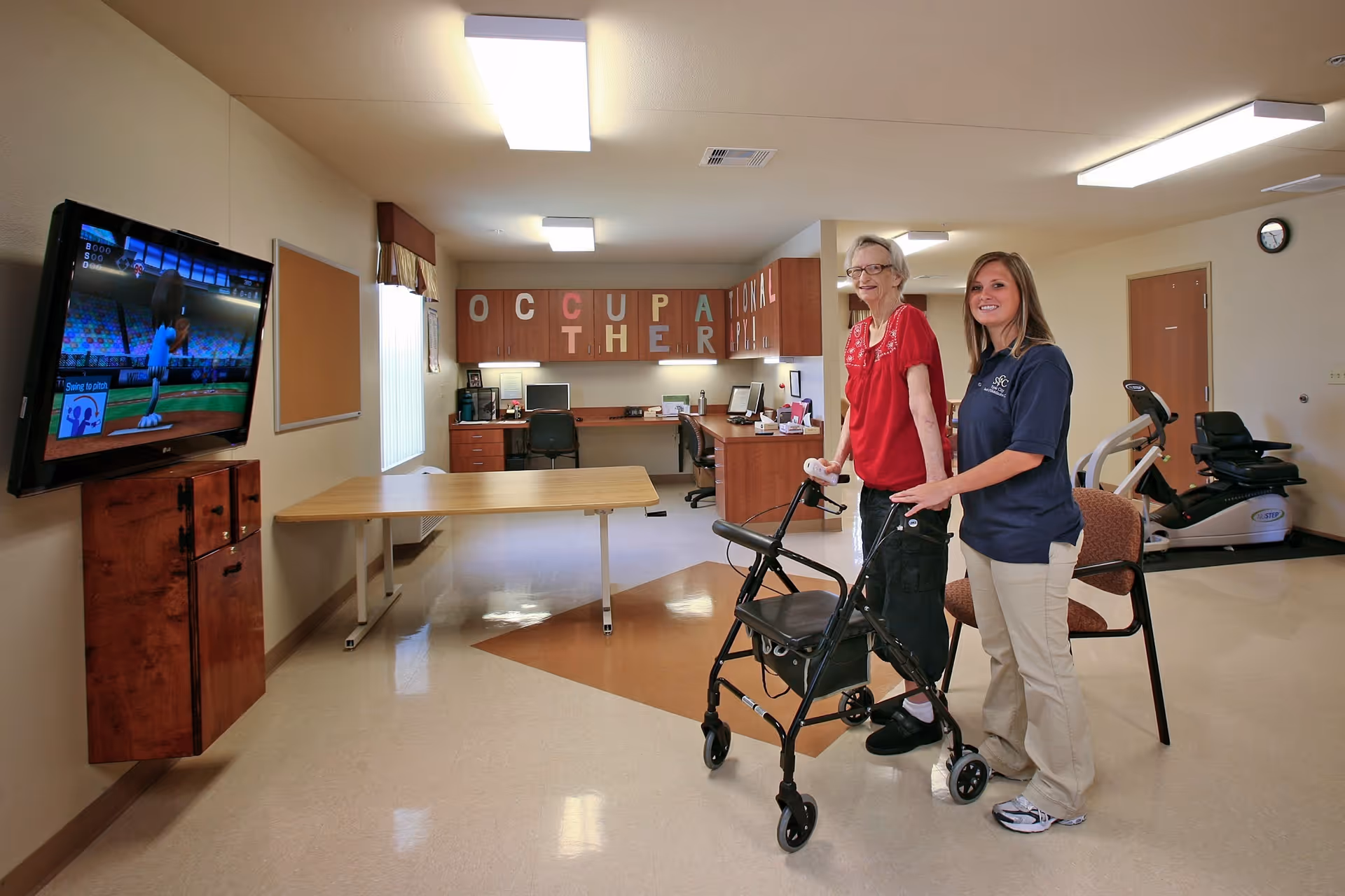 An elderly woman using a walker and a staff member stand in a bright occupational therapy room with a TV, exercise equipment, and tables.