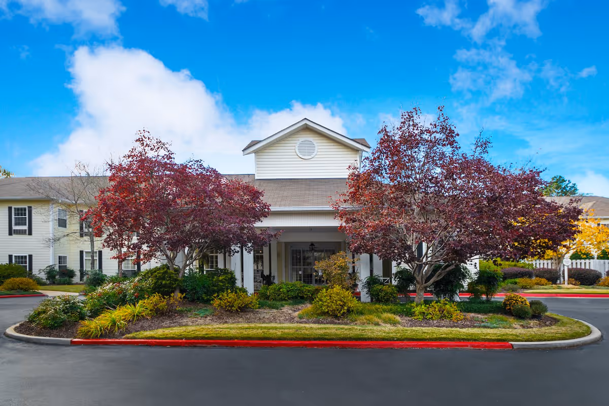 Front exterior view of Arbor Terrace Athens facility with a covered entrance, surrounded by landscaped bushes and trees with red and green foliage under a blue sky with some clouds.