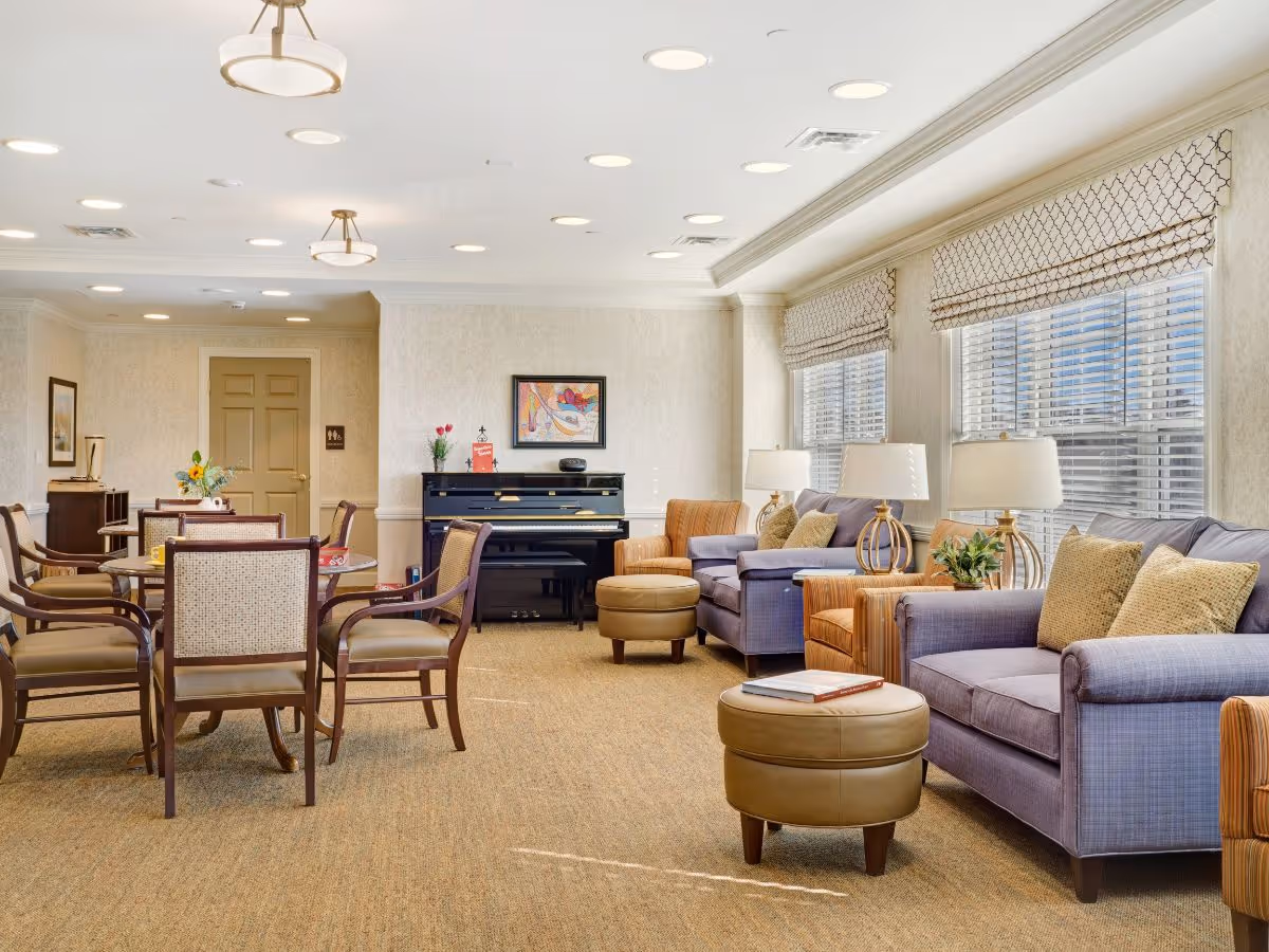 A bright and spacious common area in a senior living facility featuring comfortable seating with purple and orange armchairs, beige ottomans, and a piano against the wall. There are several tables with chairs, decorative lamps, and large windows with patterned valances allowing natural light to fill the room.