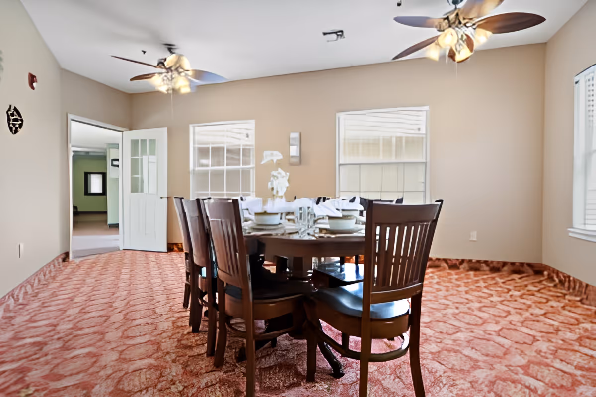 Bright dining room with a wooden table and chairs on patterned carpet beneath two ceiling fans.