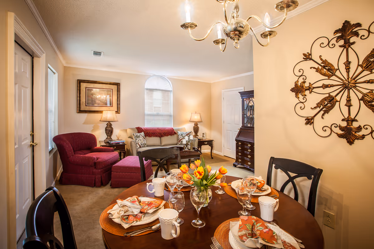 Warmly lit interior showing a set dining table in the foreground and a cozy living area with sofa and armchair in the background.
