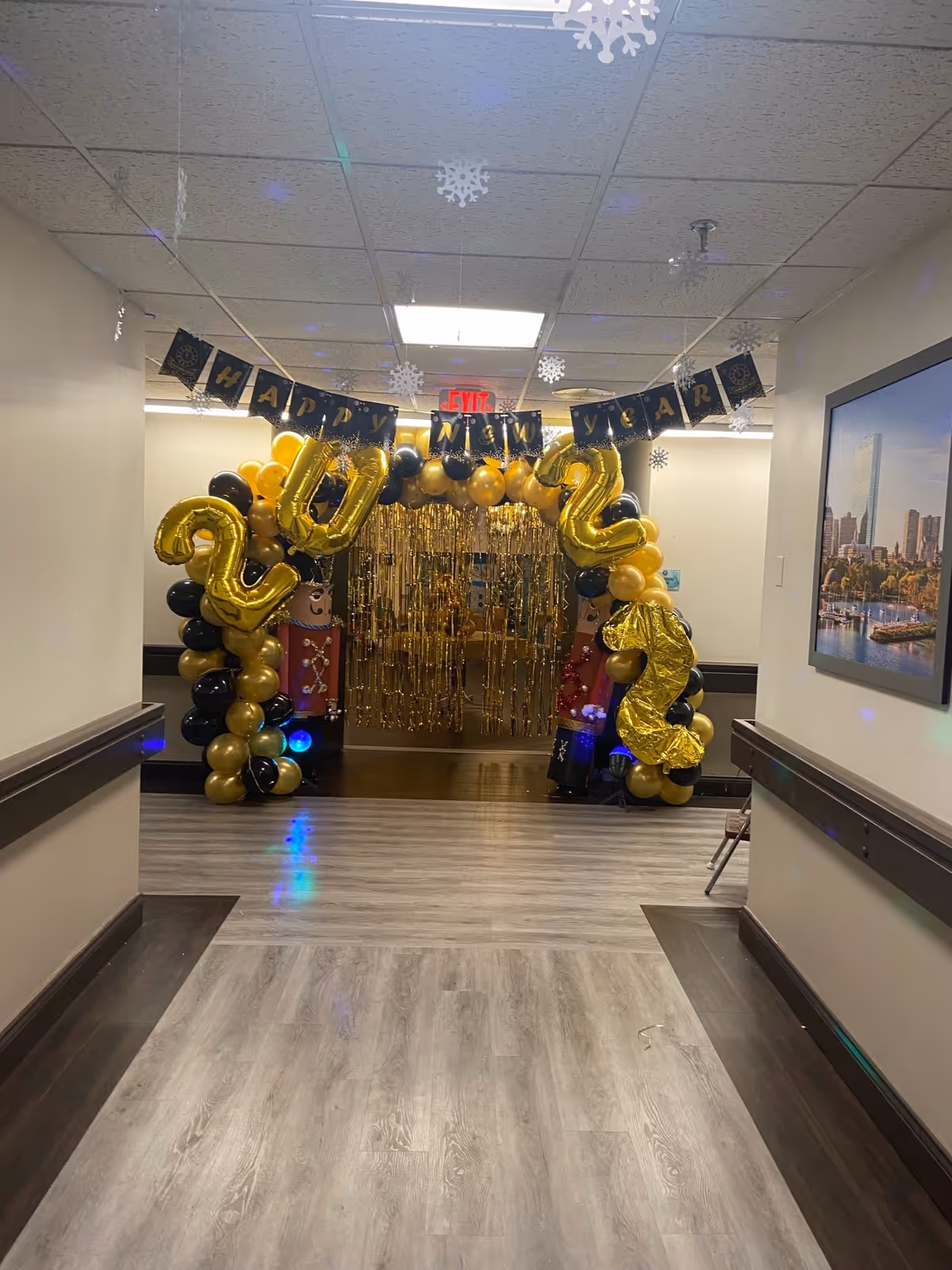 A hallway decorated for New Year's celebration with a balloon arch in black and gold colors, featuring large gold balloons forming the numbers 2023. Hanging above the arch is a banner that reads 'HAPPY NEW YEAR' with snowflake decorations hanging from the ceiling. The floor is light wood, and there is a framed picture on the right wall.