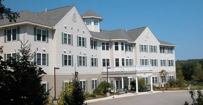 Exterior view of a large, white, multi-story senior living facility building with many windows, a covered entrance with columns, surrounded by trees and greenery under a clear blue sky.