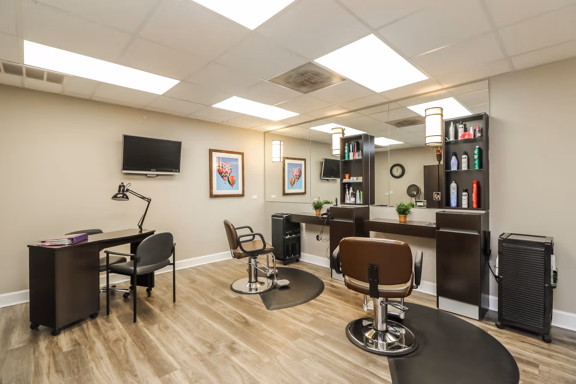 Interior of a salon room with two brown salon chairs in front of a large mirror. Shelves with hair care products are mounted on the wall next to the chairs. A small desk with two chairs and a desk lamp is positioned on the left side of the room. The floor has a wood-like finish, and the ceiling has recessed lighting panels. A wall-mounted TV and framed artwork are visible on the far wall.
