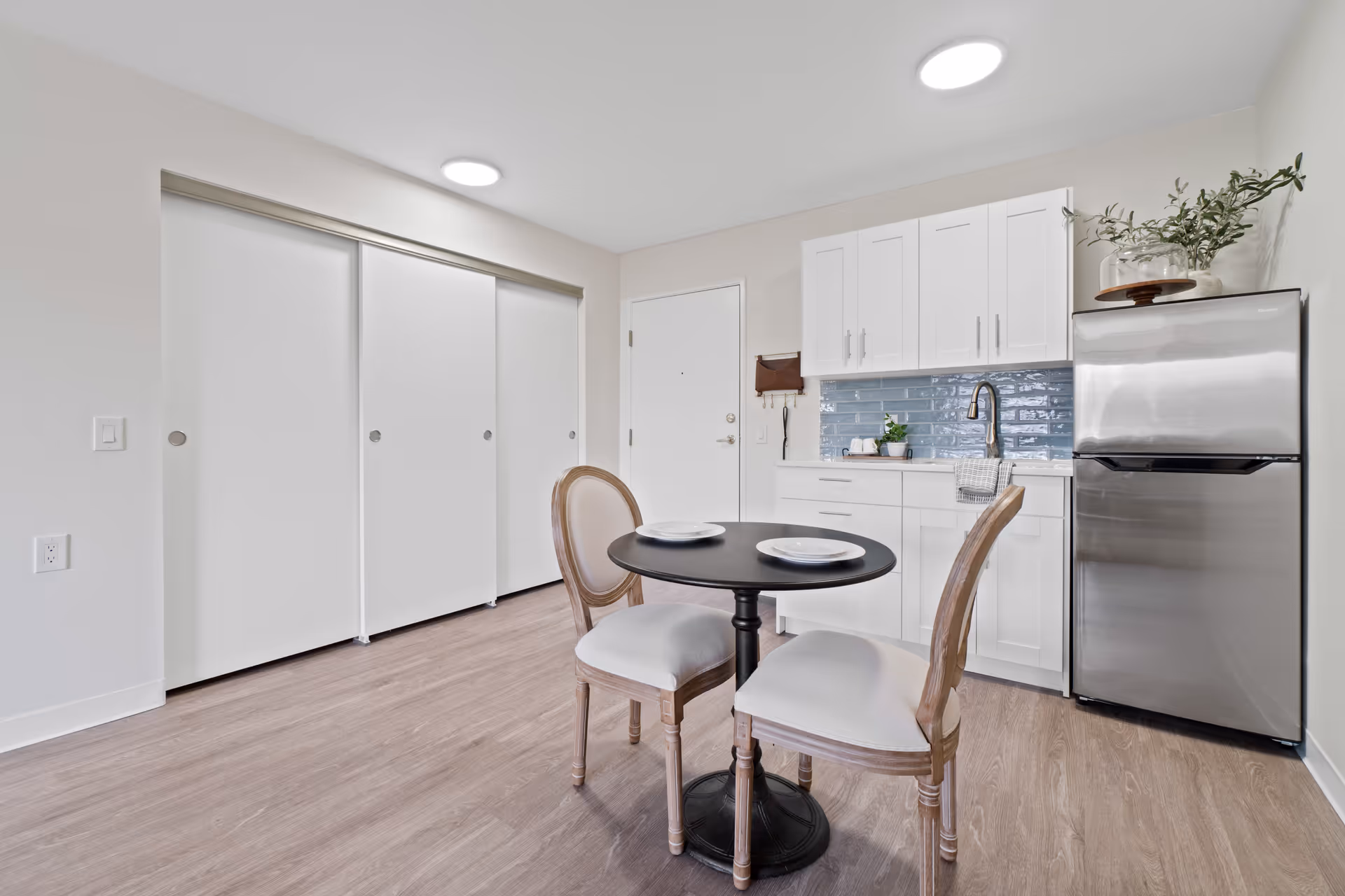 A small kitchen and dining area with white cabinets, a stainless steel refrigerator, a blue tiled backsplash, and a round black table set with two chairs and plates. There are sliding white closet doors and a white entrance door in the background.