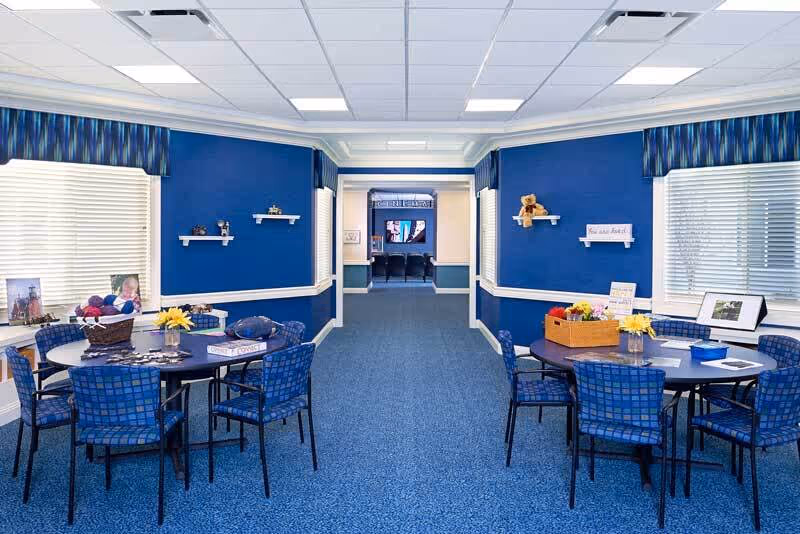A bright and clean common area with blue walls and carpet, featuring two round tables each surrounded by six blue checkered chairs. The tables have decorative items including flowers, a basket, and framed photos. The room has large windows with blinds and blue valances. At the far end, there is a hallway leading to another room with a television and chairs.