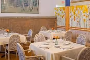 A dining room with several tables covered in white tablecloths, each set with plates, glasses, and silverware. The chairs have patterned upholstery, and there is a decorative folding screen with a floral design in the background. A framed picture hangs on the wall above a wooden panel.