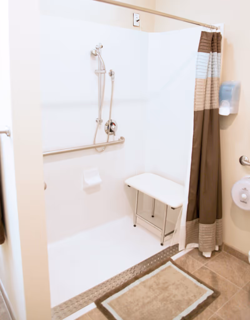 Accessible shower area with a foldable white bench, handheld showerhead, grab bar, and a brown and beige striped shower curtain. There is a beige bath mat on the tiled floor outside the shower.