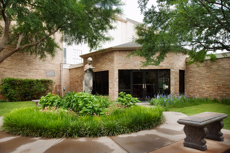 A peaceful outdoor garden area at Rolling Meadows featuring a circular flower bed with green plants and pink flowers, a classical statue in the center, a stone bench on a paved walkway, and a brick building with large windows in the background surrounded by trees.