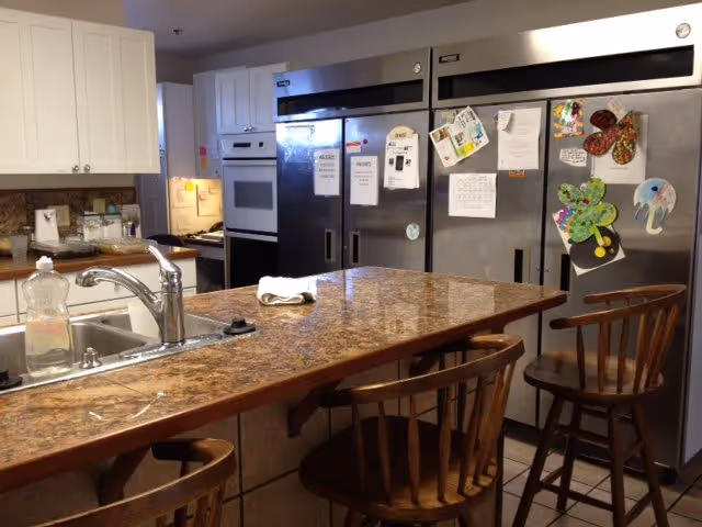 Interior view of a kitchen area featuring a granite countertop island with a sink and three wooden bar stools. In the background, there are large stainless steel refrigerators with various papers and colorful magnets attached to them. White cabinets and an oven are also visible.