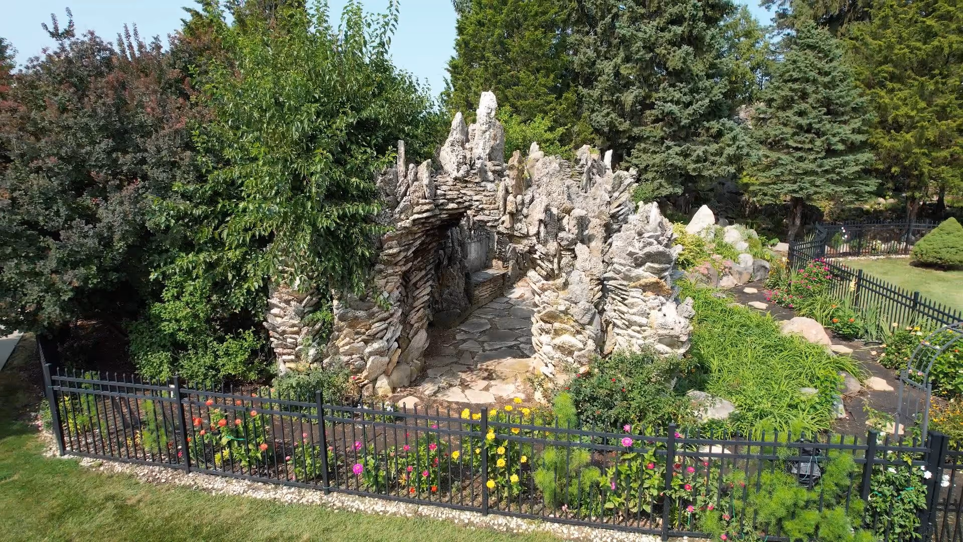 A garden area featuring a stone grotto structure surrounded by various green plants, colorful flowers, and trees. The garden is enclosed by a black metal fence and has a stone pathway leading into the grotto.