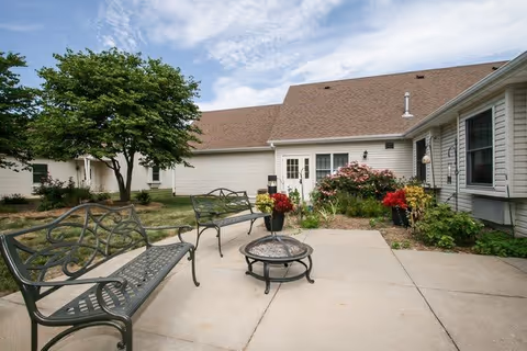 Outdoor patio area with metal benches and a fire pit, surrounded by a garden with flowers and shrubs, adjacent to a beige building with a brown roof under a partly cloudy sky.