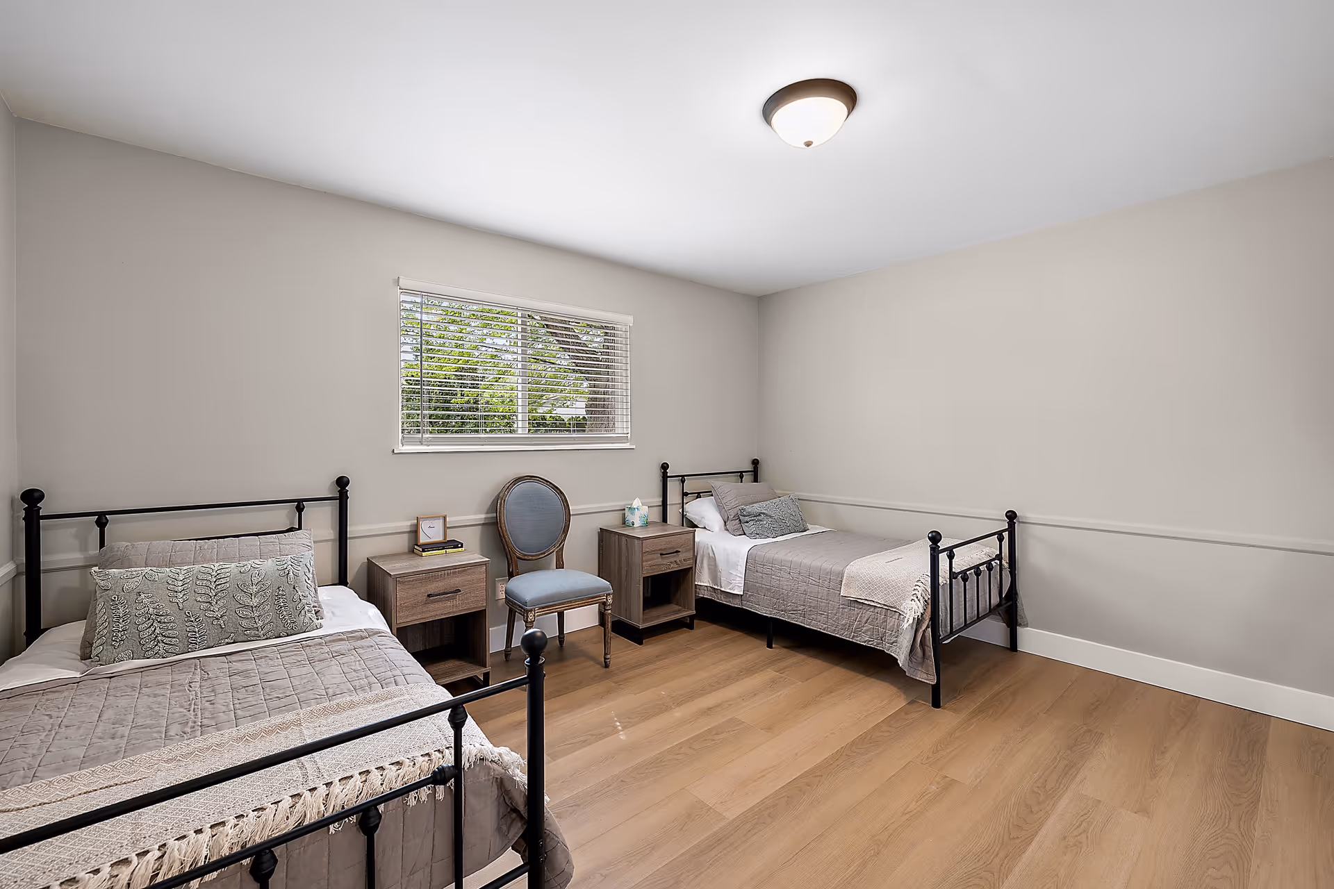 Bright bedroom with two metal-framed twin beds, two nightstands, a chair, and a window with blinds.