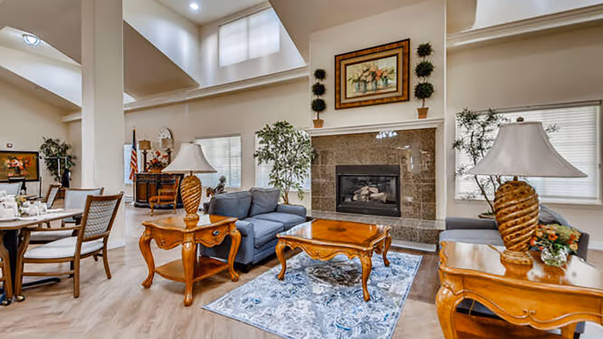 A spacious and well-lit living room area with high ceilings, featuring a blue sofa, wooden coffee and side tables with decorative lamps, a fireplace with a framed painting above it, and plants near the windows. Adjacent to the living area is a dining table with chairs.