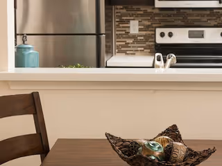 Dining table with a decorative bowl in front of a kitchen pass-through showing a refrigerator and stove.