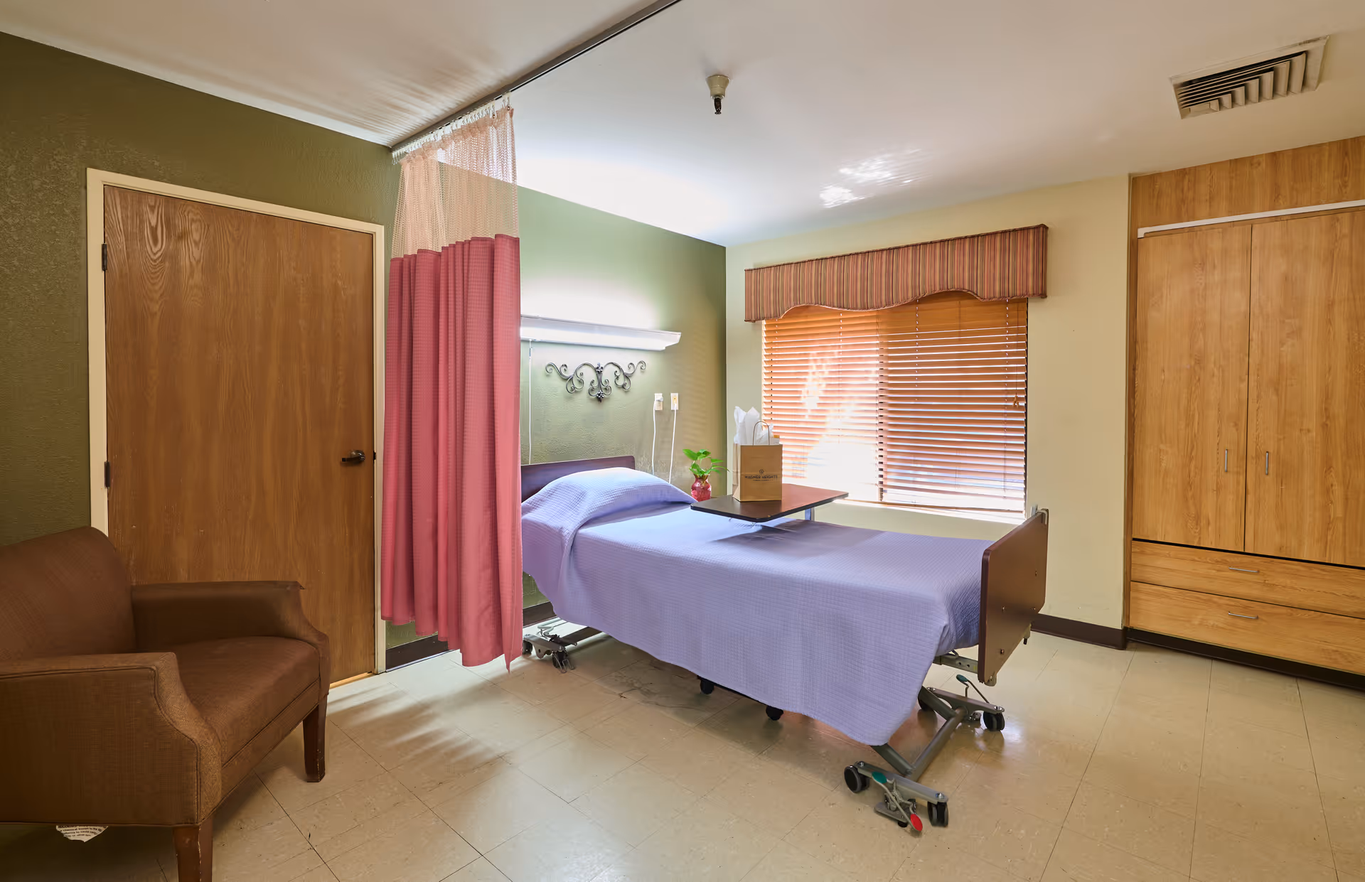 A nursing facility patient room with a hospital bed covered in a purple blanket, an overbed table, a pink privacy curtain, armchair, and wooden cabinets under a window with blinds.