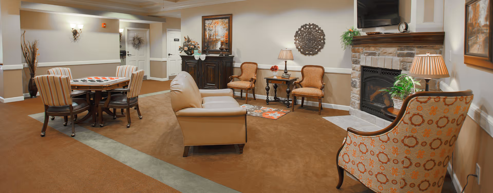 A cozy living room area in a senior living facility with a beige leather sofa, patterned armchairs, a stone fireplace, a wooden side table with a lamp, decorative wall art, and a round table with four striped chairs in the background.