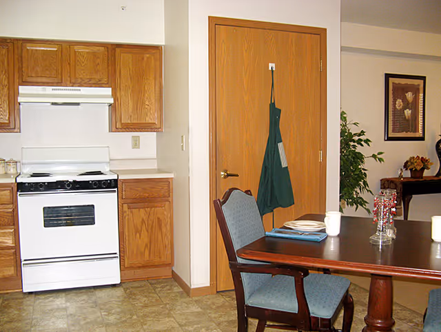 Interior view of a kitchen and dining area in a senior living facility. The kitchen features wooden cabinets, a white stove with an overhead vent, and a countertop. A wooden door with a green apron hanging on a hook is visible next to the kitchen. In the foreground, there is a wooden dining table with a blue cushioned chair, a plate, a cup, and a glass vase with decorative red beads. In the background, a framed picture and a potted plant are visible.