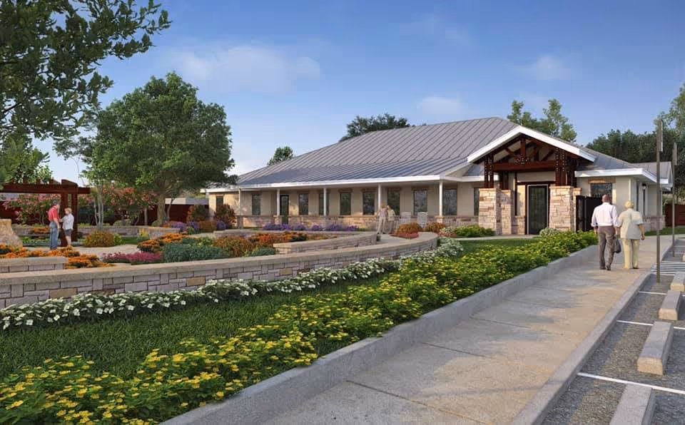 Exterior view of a single-story assisted living facility with a metal roof and stone accents. The building is surrounded by landscaped gardens with colorful flowers, shrubs, and trees. A paved walkway leads to the entrance, where two people are walking towards the building. Another two people are standing near a pergola in the garden area.