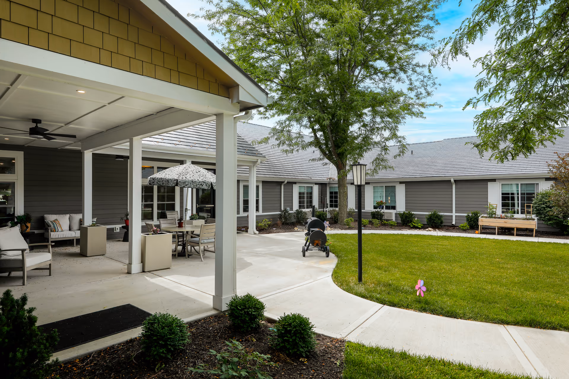 Outdoor courtyard area of Homestead Estates Assisted Living of Leawood featuring a covered patio with seating, a table with an umbrella, a grill, a walking path, green lawn, trees, and surrounding building exterior with windows.