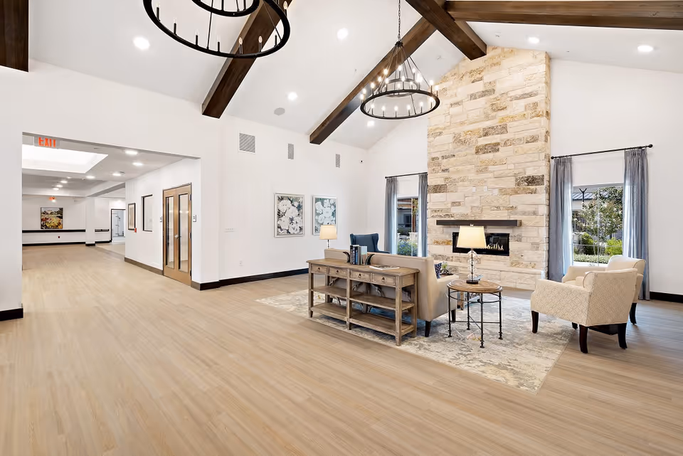A spacious and bright senior living facility common area with high vaulted ceilings featuring dark wooden beams and two large circular chandeliers. The room has light wood flooring and a stone fireplace with a modern design. There are two windows with gray curtains on either side of the fireplace. The seating area includes a beige sofa, two armchairs, a wooden console table with books, and two side tables with lamps. The walls are white with floral artwork, and a hallway with double glass doors is visible on the left side.