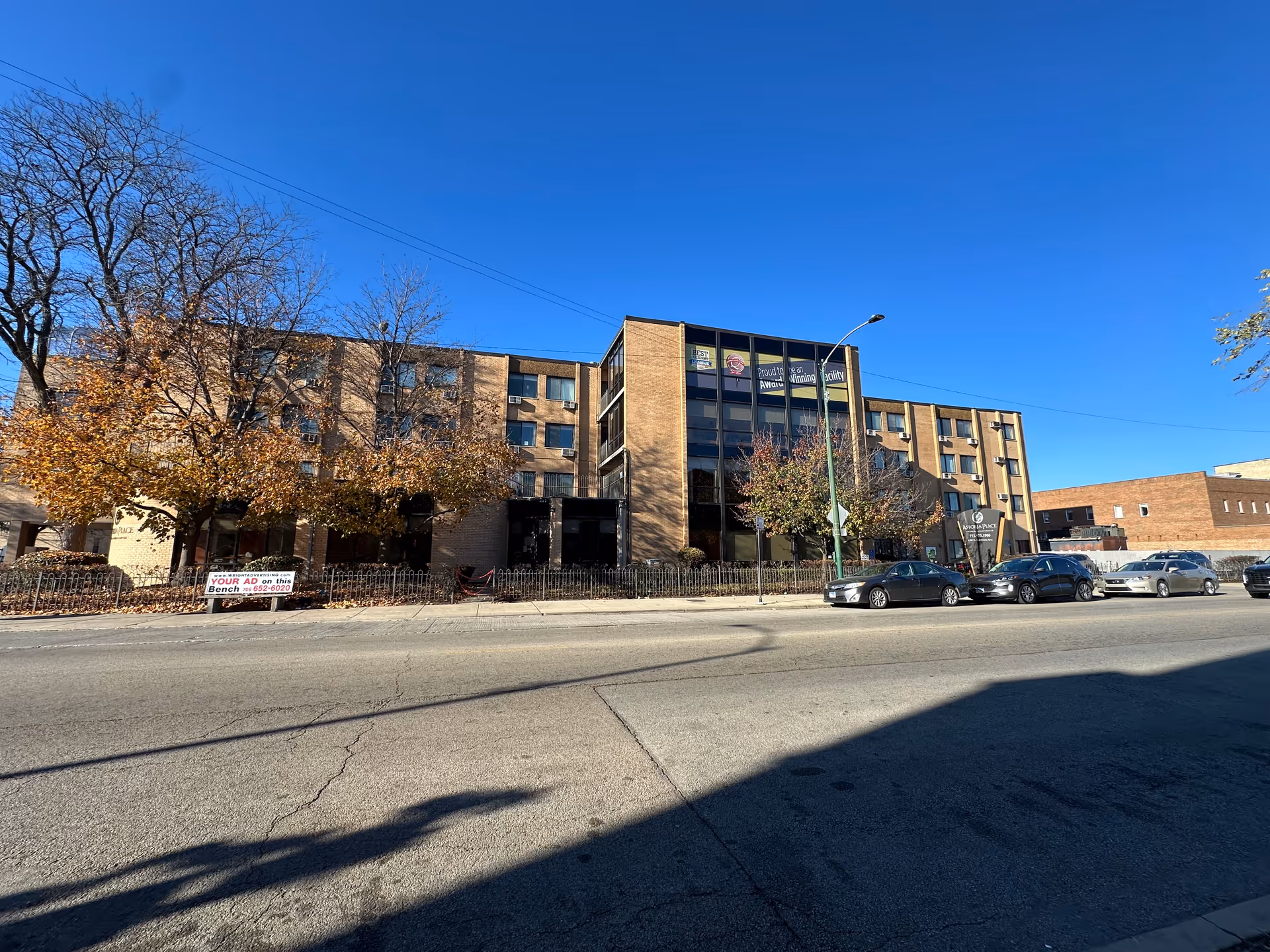 Exterior view of a multi-story brick building with large windows and several trees with autumn leaves in front. Several cars are parked along the street in front of the building under a clear blue sky.