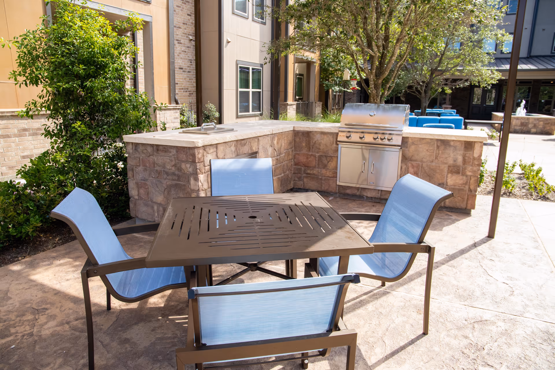 Outdoor patio area with a square metal table surrounded by four blue mesh chairs. In the background, there is a built-in stone grill and sink, with trees and shrubs nearby. The setting is sunny and appears to be part of a residential or senior living facility courtyard.