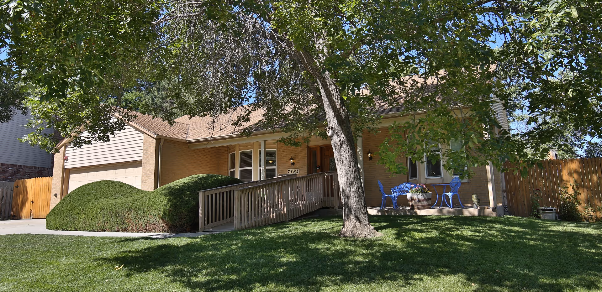 A single-story brick house with a sloped roof, a wooden ramp leading to the front door, and a small patio area with blue chairs and a table. The house is surrounded by green grass, a large tree in the front yard, and neatly trimmed bushes. A wooden fence encloses the backyard.