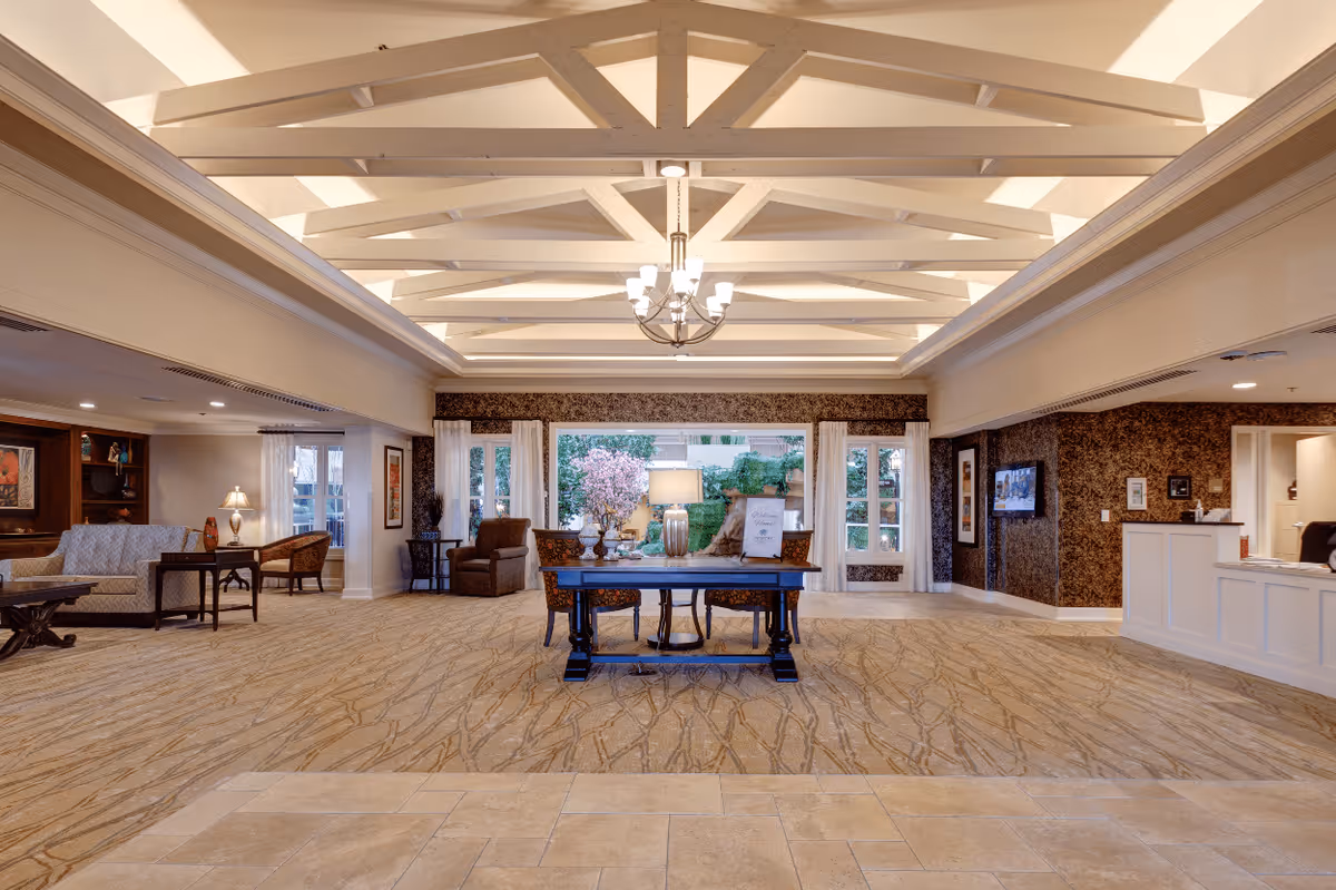 Spacious senior living facility common area with a high ceiling featuring exposed beams and a chandelier. The room has a patterned carpet and tiled floor near the entrance. A wooden table with decorative items and a lamp is centered in the room. Comfortable seating areas with armchairs and sofas are arranged along the sides. Large windows and glass doors at the far end provide a view of greenery outside. The reception desk is visible on the right side of the image.