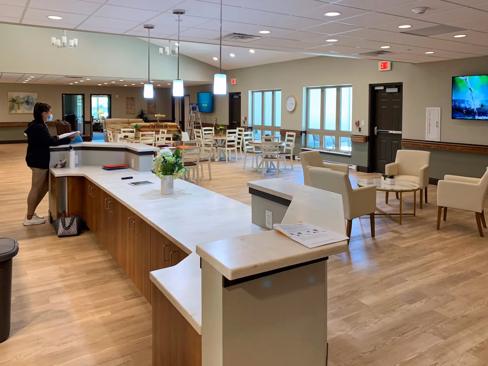 Interior view of a senior living facility common area with a reception desk in the foreground, a woman wearing a mask standing behind the desk, several tables and chairs arranged in the middle, and a seating area with armchairs and a round table. The room has large windows, pendant lights, and a wall-mounted TV.