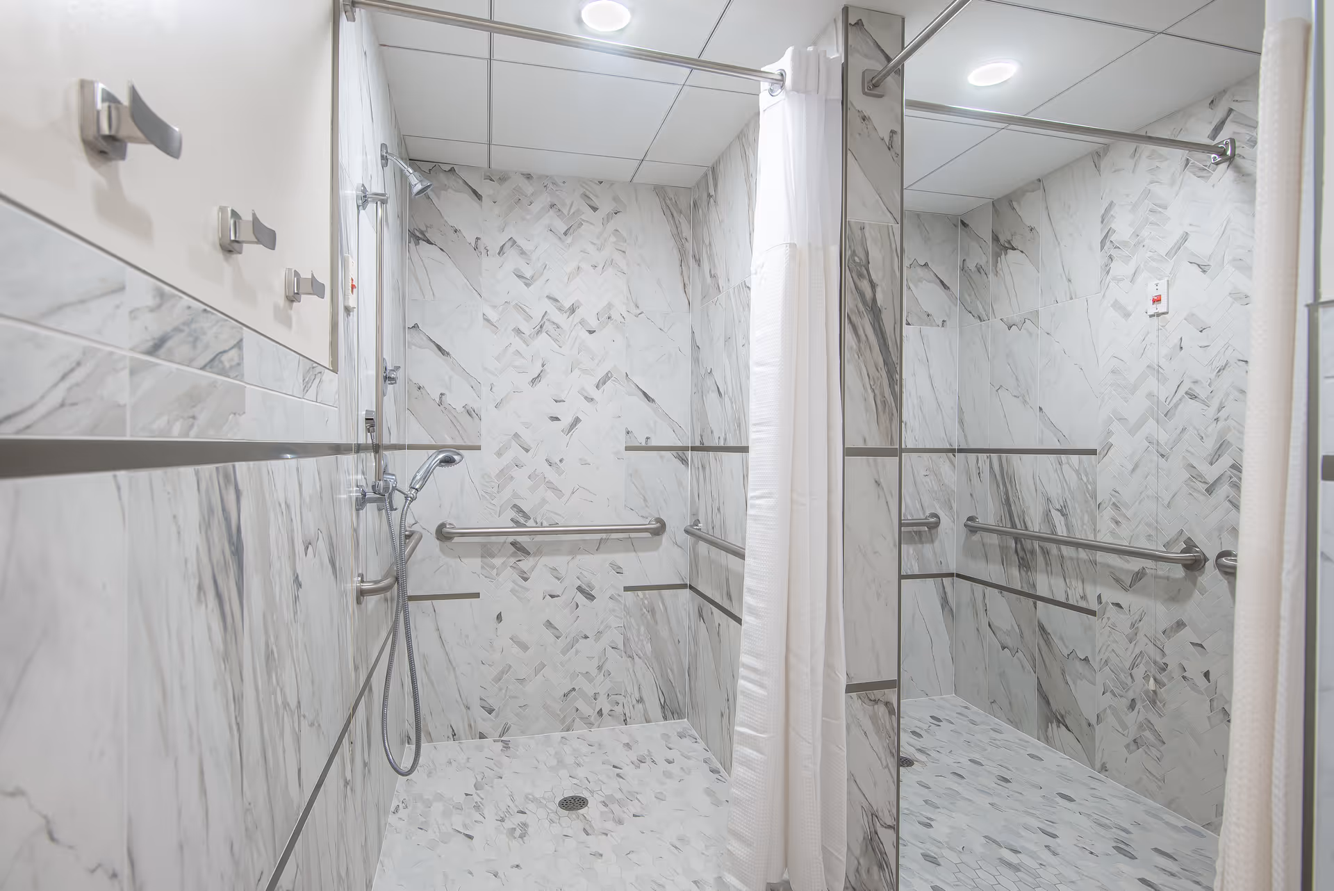 A spacious, modern shower area with marble-patterned tiles on the walls and floor. The shower features a handheld showerhead, grab bars for safety, and a white shower curtain. The ceiling has recessed lighting, and there is a large mirror reflecting the shower space.