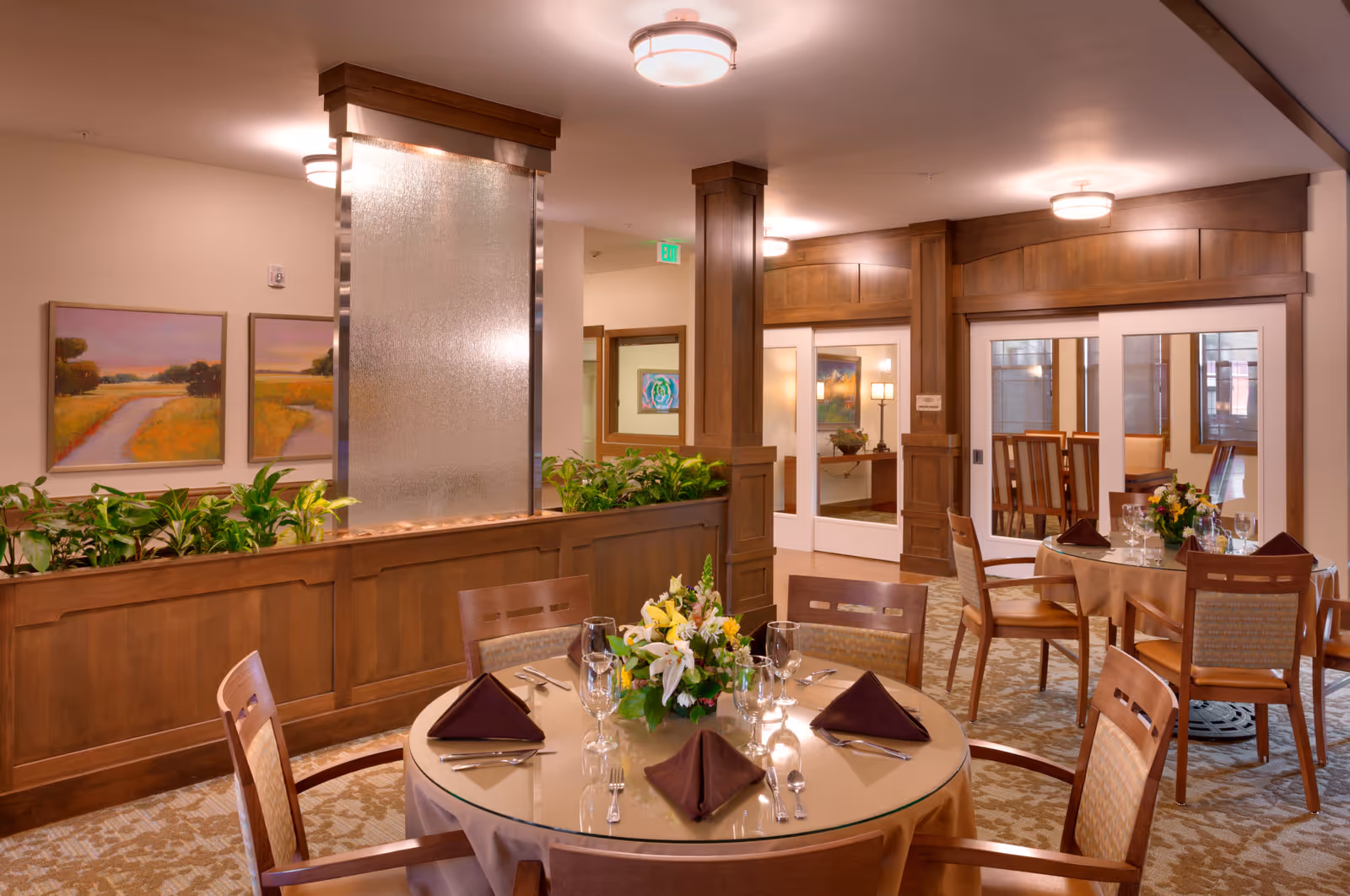 Well-appointed dining room with round tables set for guests, wooden paneling, plants, and artwork on the walls.