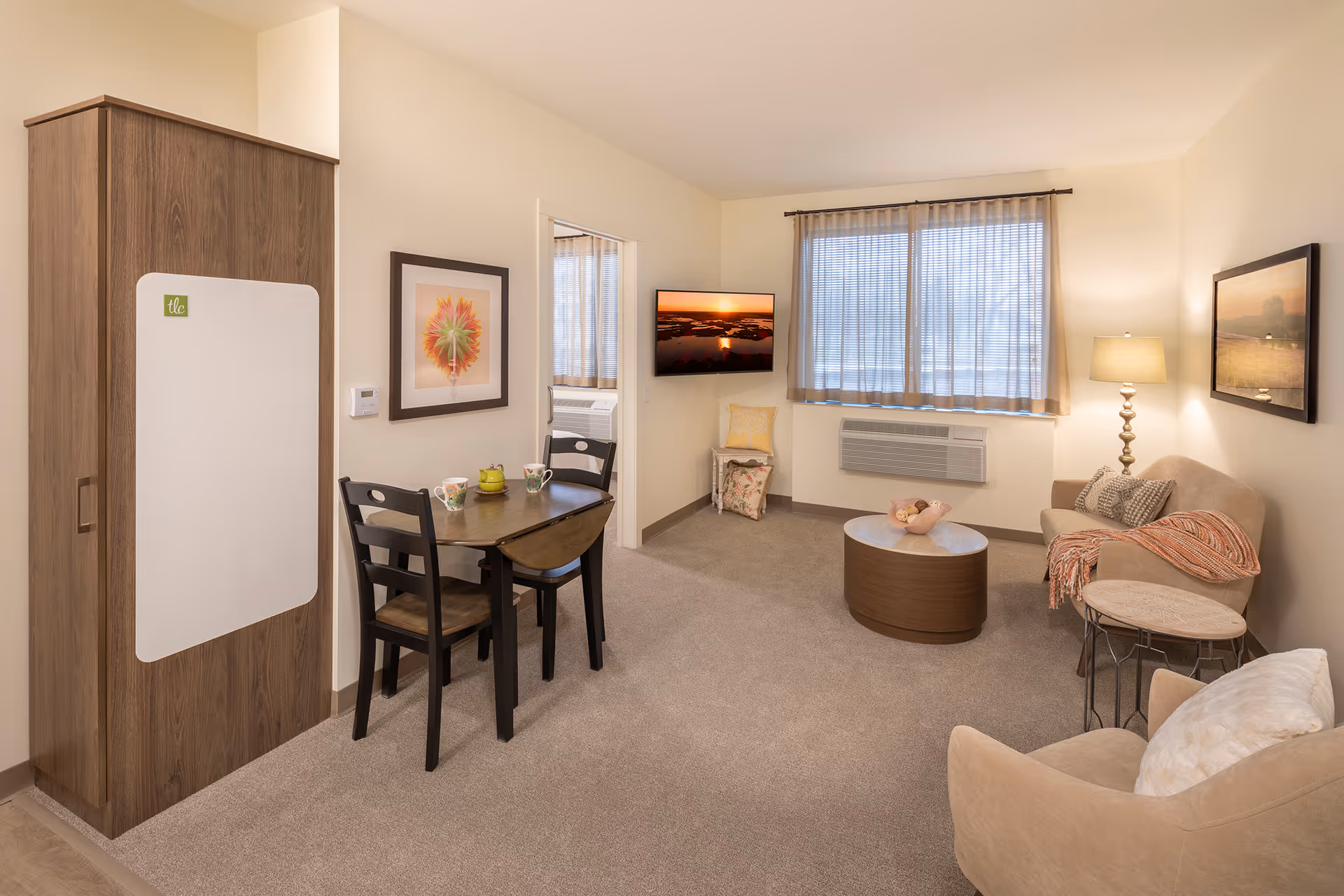 Bright furnished living room with a small dining table, sofa, armchairs, coffee table and wall-mounted TV in a senior living apartment.