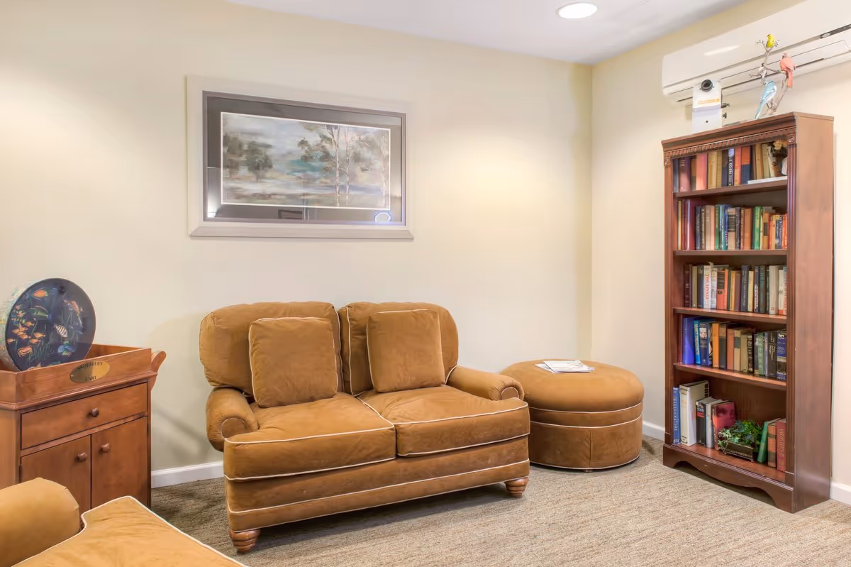A cozy sitting area with a brown loveseat and matching ottoman on a carpeted floor. There is a wooden bookshelf filled with books on the right side and a small wooden cabinet with a decorative plate on the left. A framed landscape painting hangs on the beige wall above the loveseat. An air conditioning unit is mounted on the wall above the bookshelf.