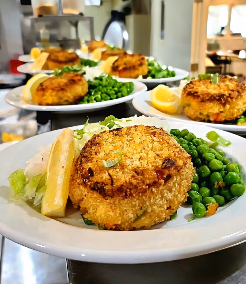 Close-up view of multiple white plates each containing a golden-brown crab cake, a lemon wedge, green peas, and a small portion of shredded lettuce, arranged in a kitchen or food service area.