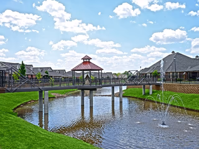 A scenic outdoor view of a pond with a water fountain in the foreground, a pedestrian bridge with a red-roofed gazebo in the center crossing over the pond, and residential buildings with pitched roofs in the background under a partly cloudy blue sky.