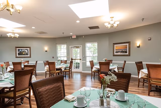 A dining room with several round tables covered with green and white tablecloths. Each table is set with cups, glasses, napkins, and small flower arrangements. The room has wooden chairs, light gray walls with framed paintings, and ceiling lights including a chandelier. There are windows and a glass door letting in natural light.