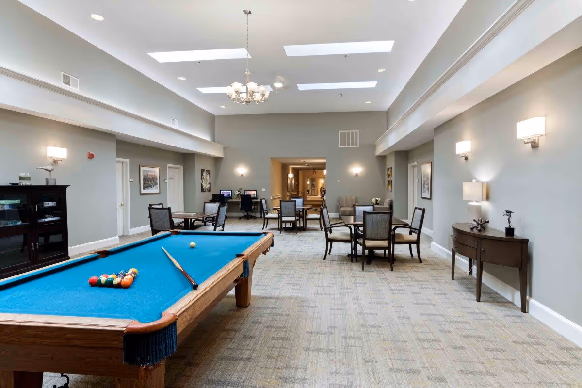 A spacious common area with a pool table in the foreground, several tables and chairs arranged for seating, two computer stations against the far wall, soft lighting from wall sconces and ceiling fixtures, and neutral-colored walls and carpet.