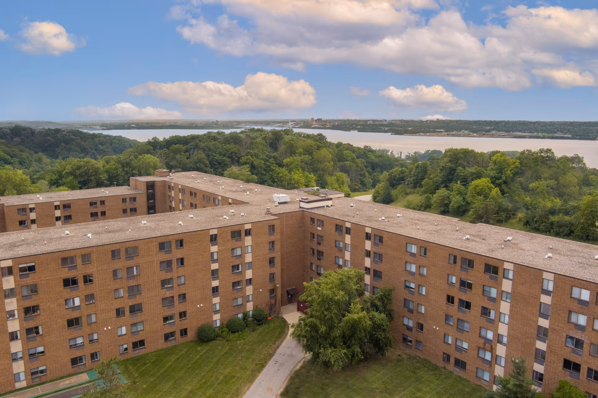 Aerial view of a large, multi-story brick senior living facility surrounded by green trees and grass, with a river and forested area in the background under a partly cloudy sky.