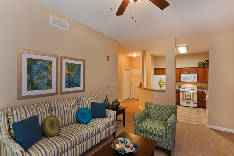 A cozy living room area with a striped sofa adorned with blue and green pillows, a patterned armchair, and a wooden coffee table with decorative items. Two abstract paintings hang on the beige wall above the sofa. In the background, there is a kitchen with wooden cabinets, a white refrigerator, stove, and microwave. The space is warmly lit with ceiling lights and a ceiling fan with wooden blades.
