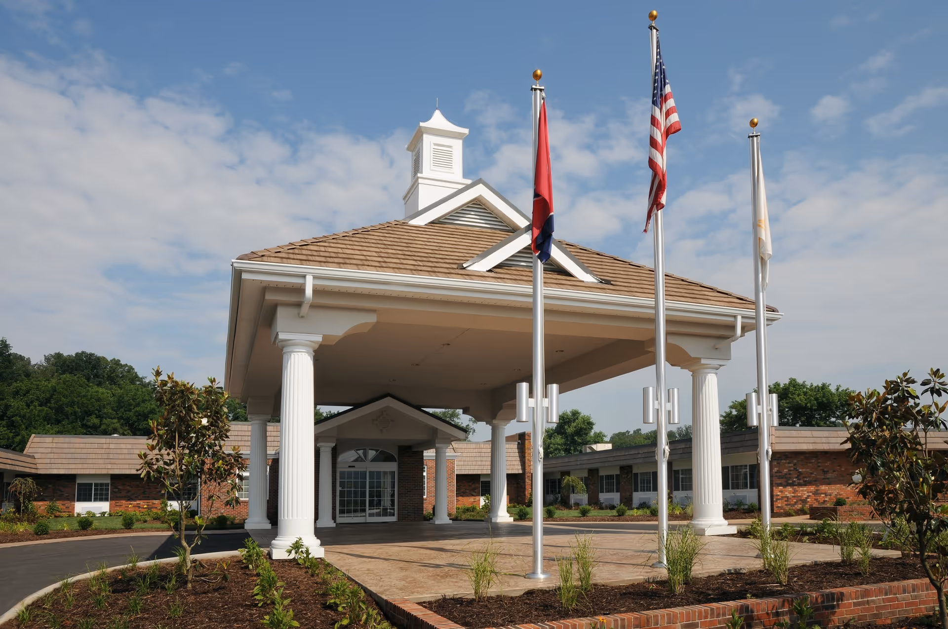 Entrance of a senior living facility with a covered driveway supported by white columns, three flagpoles with flags, and a brick building in the background under a partly cloudy sky.