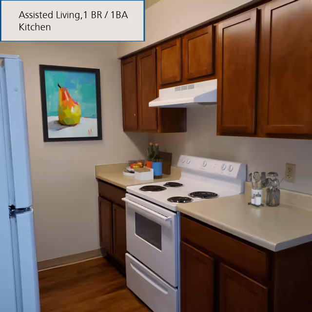 A kitchen area in an assisted living facility featuring wooden cabinets, a white electric stove with four burners, a white refrigerator, and a countertop with utensils and decorative items. A colorful framed painting of a pear hangs on the wall.