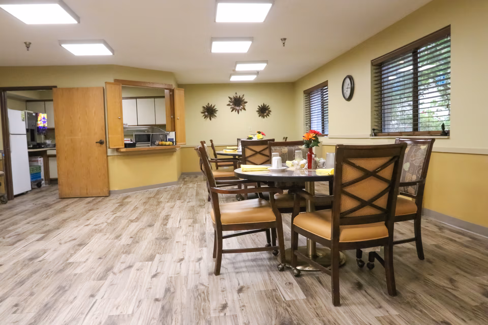 A dining room in a senior living facility with round tables and chairs arranged for meals. The tables have place settings with cups, plates, and napkins, and there are flowers in vases as centerpieces. The room has wood-patterned flooring, yellow walls, windows with blinds, and a clock on the wall. A serving window and a door leading to a kitchen area are visible in the background.